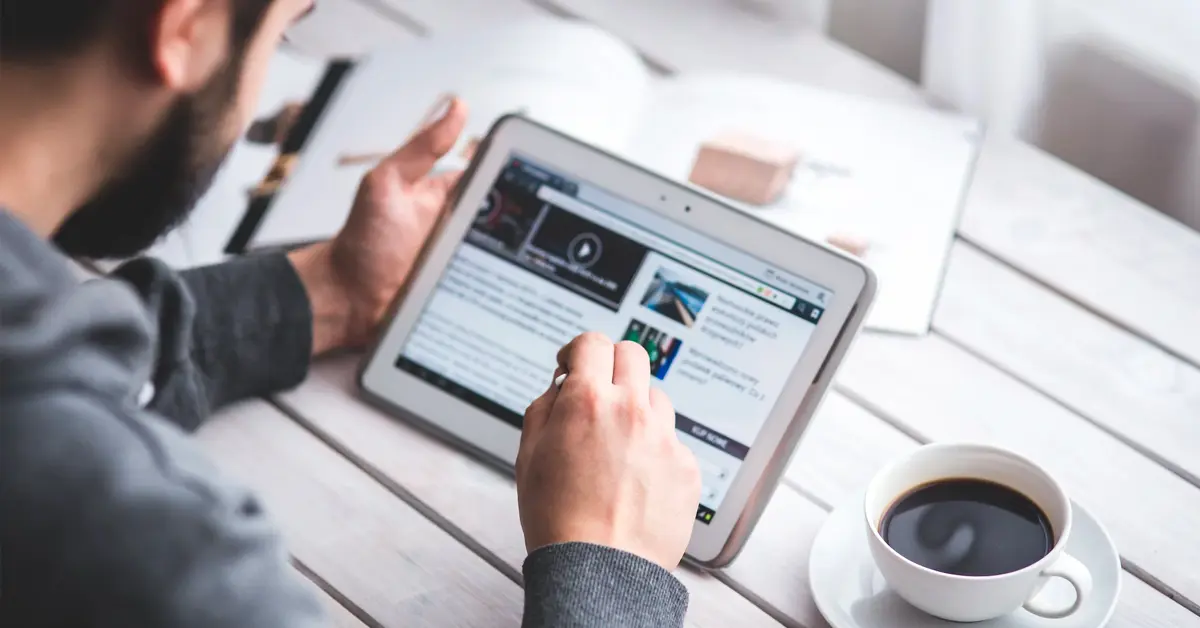 A man using the tablet while drinking a coffee. In the background, there's an open book.